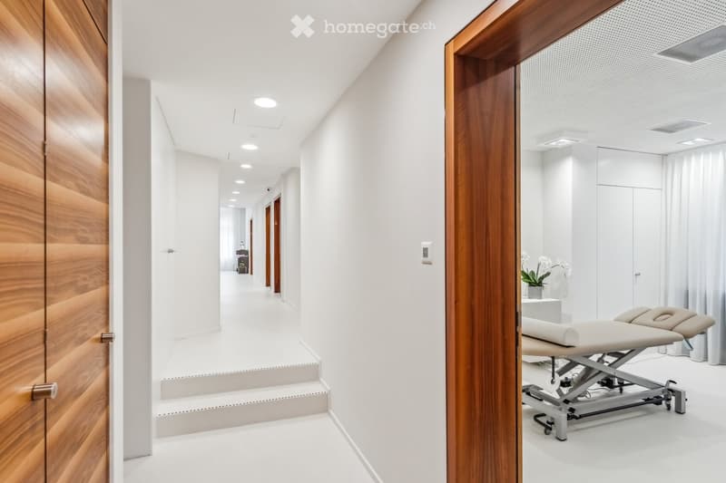 Modern, clean hallway with white walls, recessed lighting, and wooden paneling on one side. An open doorway leads to a treatment room with a massage table, white curtains, and a small plant on a stand.