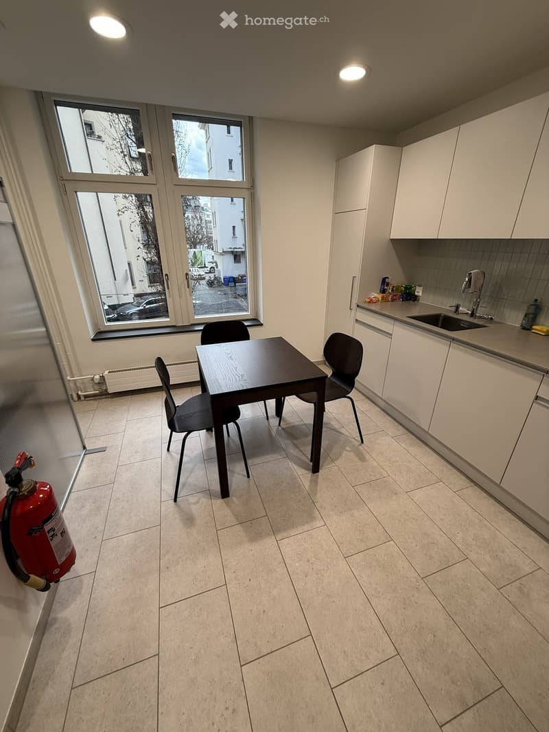 Modern kitchen with white cabinets, a small dining table with four chairs, and a sink area with a faucet. The room has large windows offering a view of the street outside.