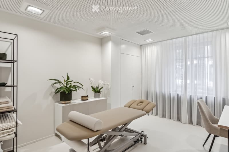 Modern, minimalist room with a massage table, a chair, a small table with a plant and decorative items, a shelf with towels, and large windows with sheer curtains.