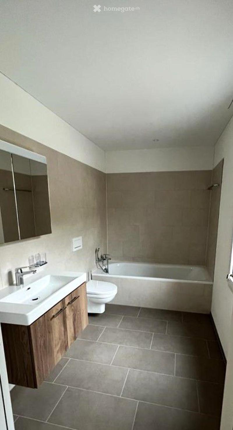 Modern bathroom with a white sink, wooden cabinet, mirror, toilet, and bathtub. The walls are tiled in a neutral color, and the floor is covered with dark gray tiles.