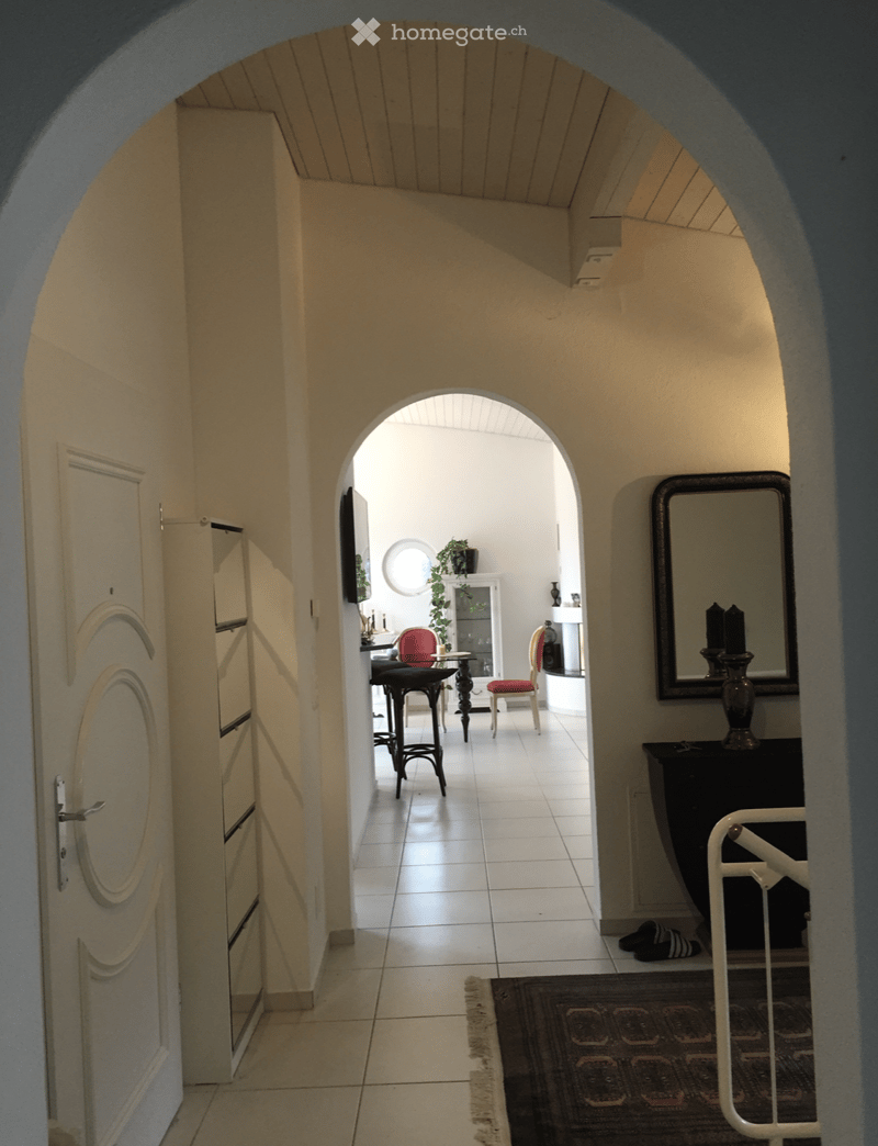 A well-lit hallway with arched doorways leading to another room, featuring tiled flooring, a small table with chairs, and a mirror on the wall.