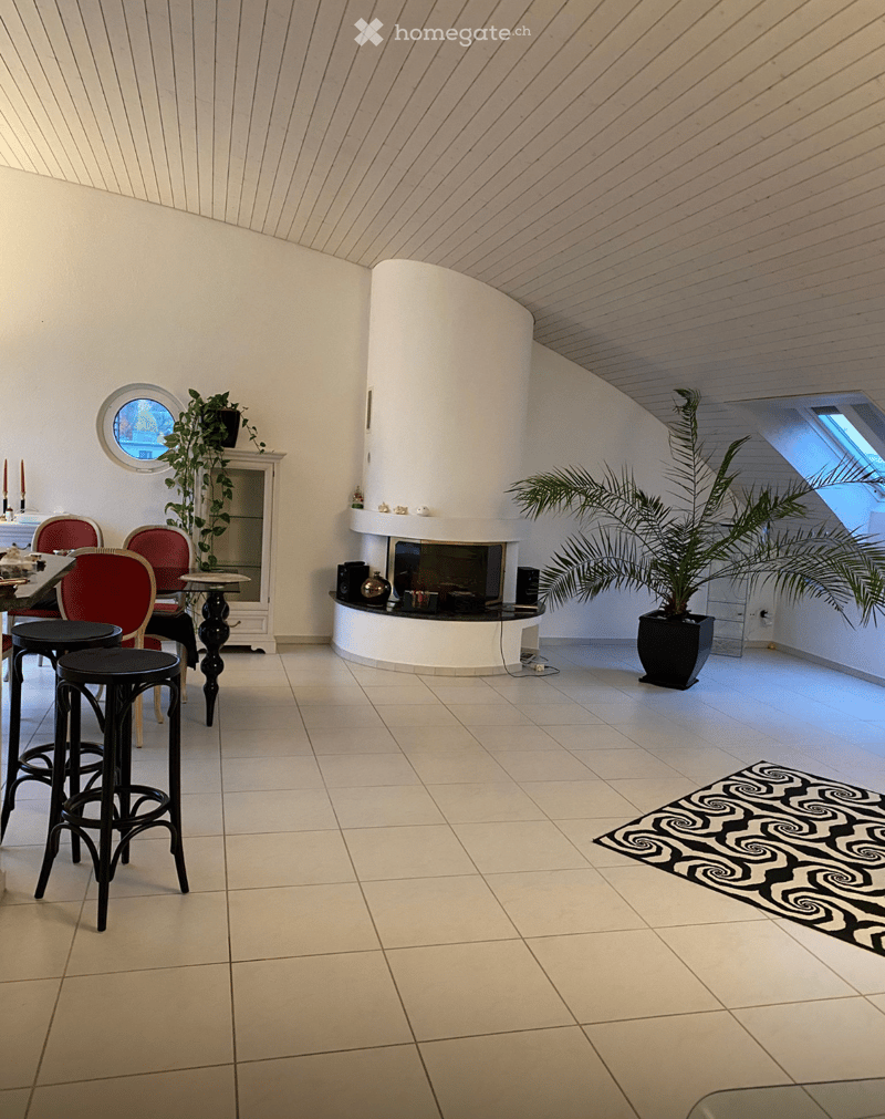 Bright living room with white walls, tiled floor, and a curved white fireplace. Features include a round table with red chairs, black stools, a large potted plant, and a decorative black and white rug.