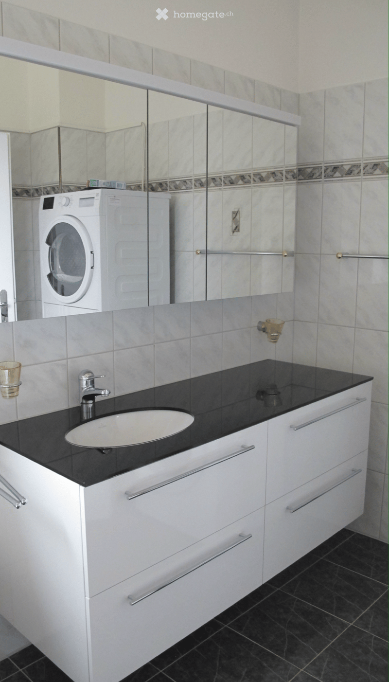 Modern bathroom with white cabinets, black countertop, sink, faucet, and a washing machine in the background.