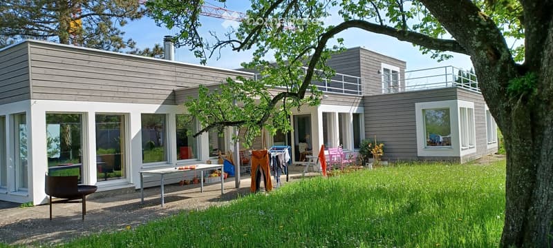 Modern two-story house with grey wooden siding, large windows, and a balcony on the upper level. The surrounding area includes a grassy lawn, a tree, and outdoor furniture near the entrance.