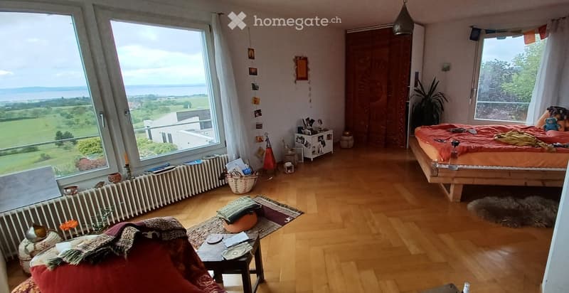 Bright bedroom with wooden flooring, large windows offering a scenic view, a bed with orange bedding, a small table with a chair, a plant, and decorative items.
