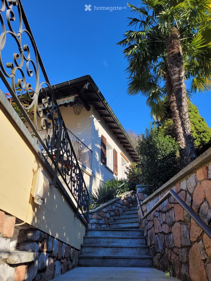 A two-story residential building with a stone staircase leading up to a balcony, featuring ornate metal railings and a palm tree nearby.