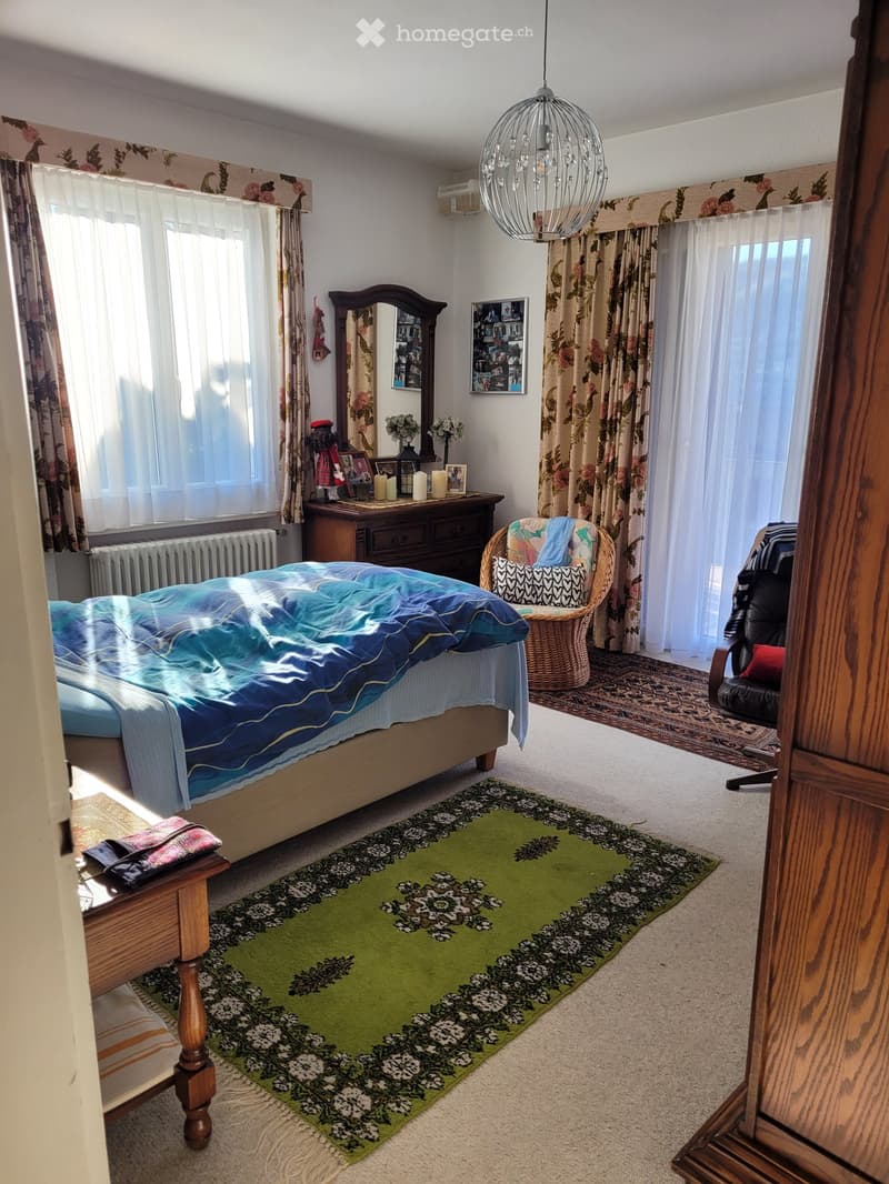 Cozy bedroom with a large bed, nightstand, wicker chair, mirror, and decorative items on a dresser. The room features patterned rugs and floral curtains.