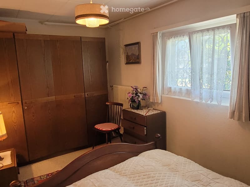 Cozy bedroom with a wooden bed, wardrobe, small table with a flower vase, chair, and a picture on the wall. Natural light filters through lace curtains.