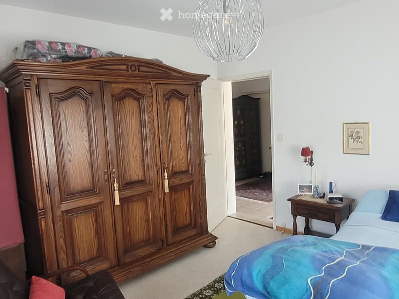 A well-furnished bedroom with a large wooden wardrobe, a neatly made bed with blue bedding, a nightstand with a lamp, and a picture frame on the wall.