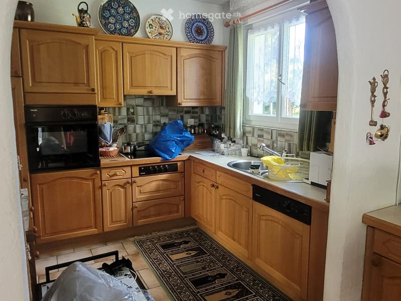 A cozy kitchen with wooden cabinets, a black oven, a sink with a faucet, and a dishwasher. The counter has various items including a teapot, plates, and a basket. A patterned rug is on the floor.