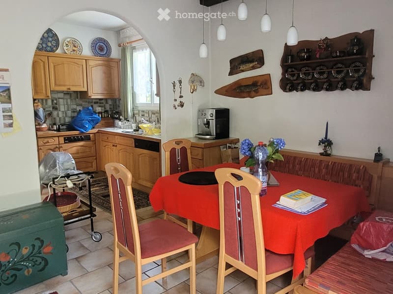 A cozy kitchen and dining area with wooden cabinets, a red tablecloth-covered dining table, four chairs, a sofa, and a coffee maker on the counter.