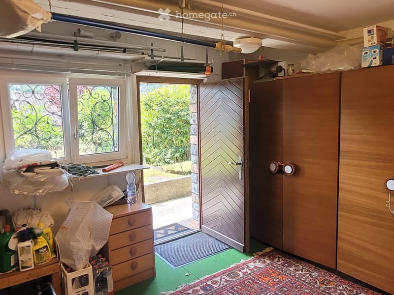 A small, cluttered utility or storage room with a wooden cabinet, a small wooden drawer unit, shelves holding various items, cleaning supplies, and a partially open door leading outside.