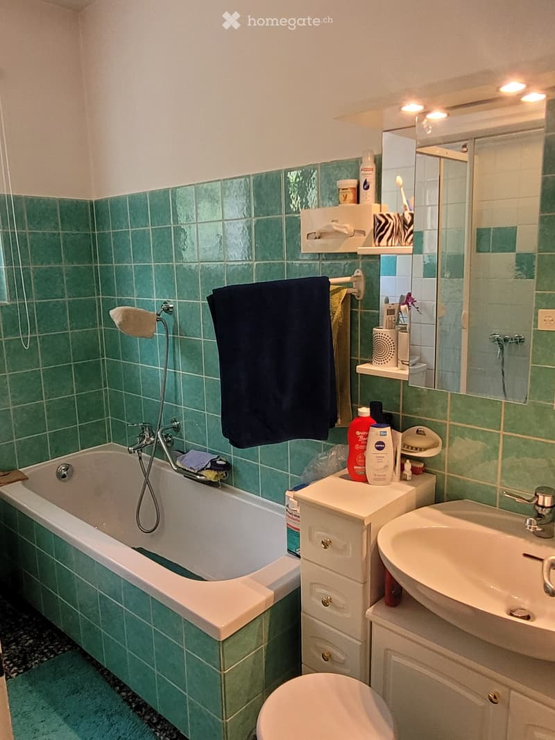 Bathroom with green tiled walls, bathtub, sink, toilet, mirror, and shelves holding toiletries and towels.