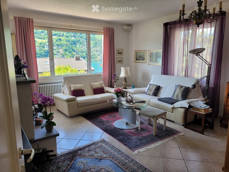 Bright living room with large windows offering a scenic view, featuring a white sectional sofa, glass coffee table, floor lamp, and decorative items.