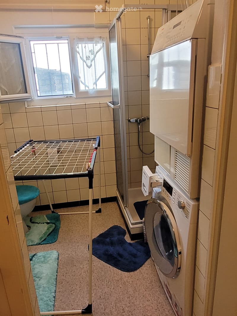 Bathroom with a washing machine, drying rack, toilet, shower stall, and bathmats.