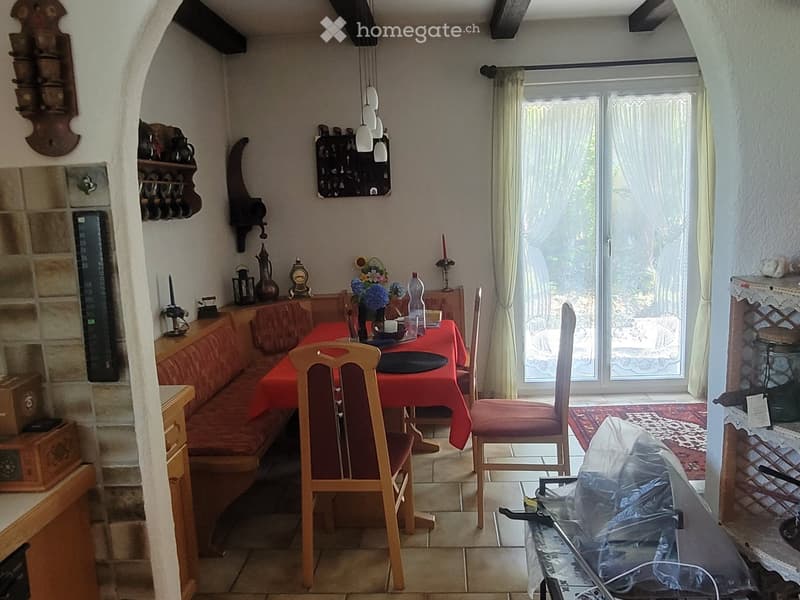 Cozy dining area with a red tablecloth, wooden chairs, a sofa, and decorative items including a flower vase, candle, and table lamp. Large windows with curtains lead to an outdoor view.