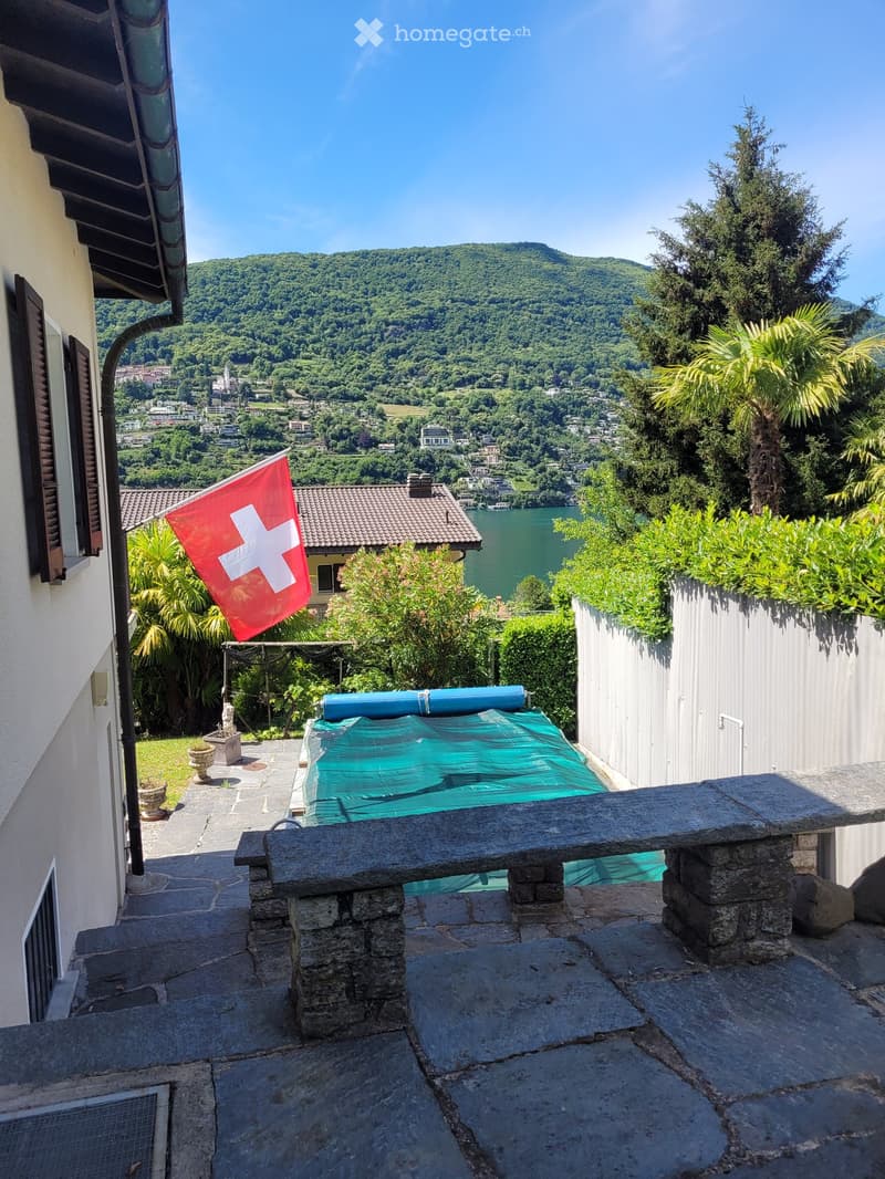Balcony with stone bench, covered swimming pool, Swiss flag, and scenic view of a lake and mountains.
