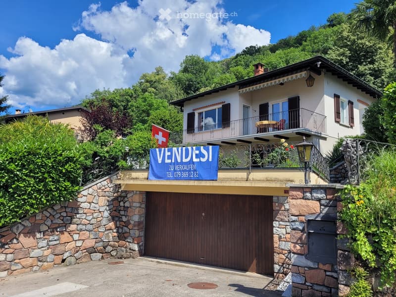 Two-story house with a garage, balcony, and Swiss flag, surrounded by greenery and trees.