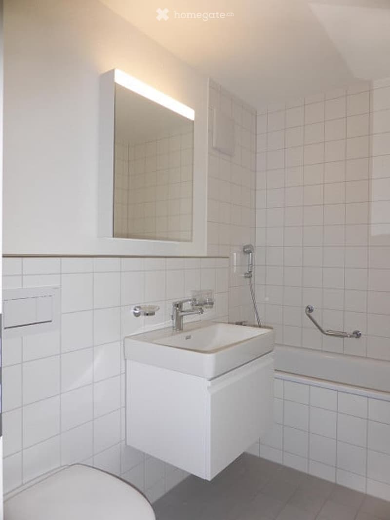 Modern bathroom with white tiles, a white sink with a modern faucet, a large mirror with built-in lighting, and a bathtub with a handrail.