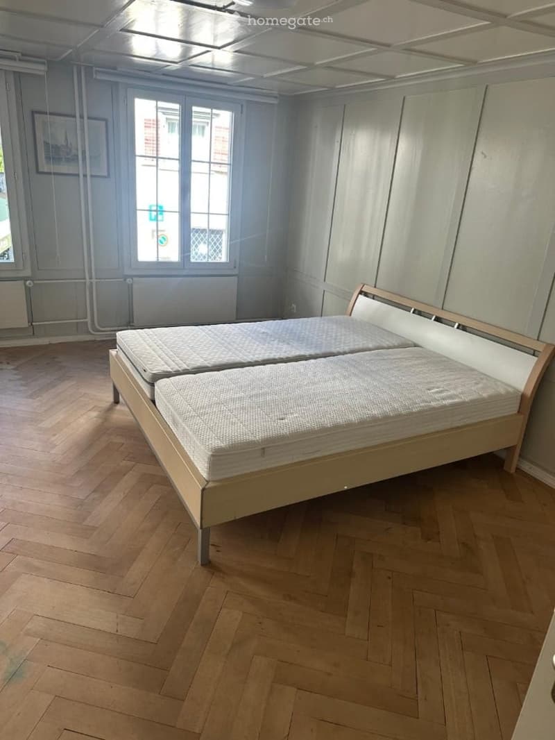 Bright bedroom with wooden floor, large window, and a single bed with white mattress and headboard.