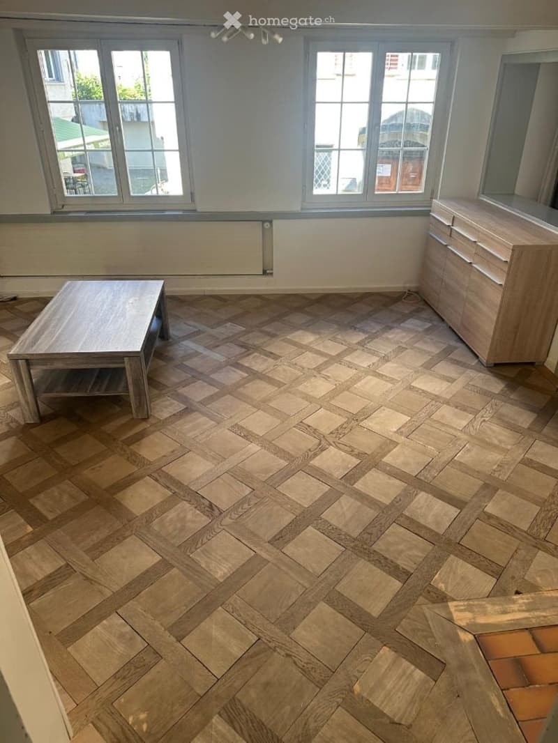 The room features a herringbone-patterned wooden floor, large windows, a simple wooden table, and a light wooden cabinet with drawers.