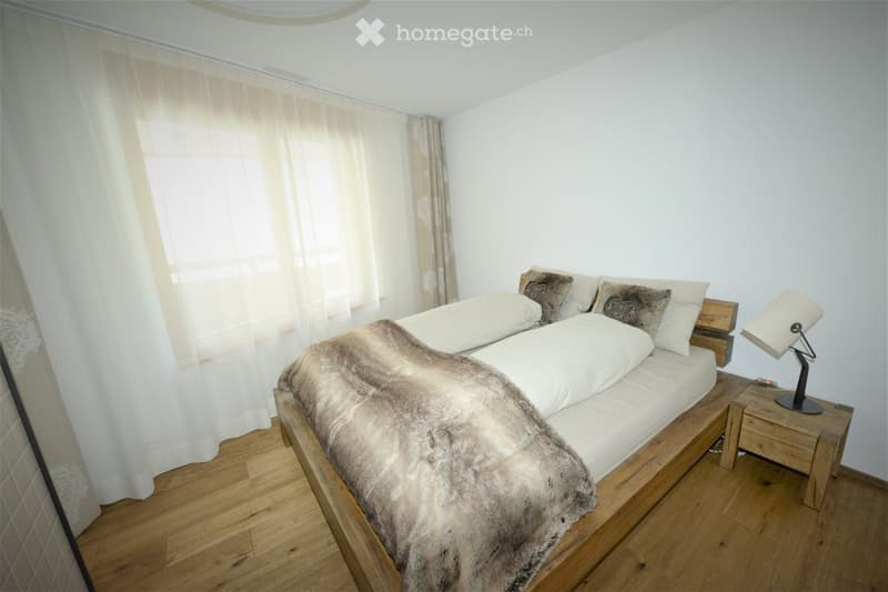 Cozy bedroom with wooden flooring, a single bed with a fur throw and pillows, a nightstand with a lamp, and light-colored curtains.
