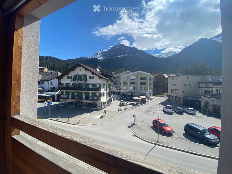 Balcony view overlooking a town square with buildings, parked cars, and distant mountains.