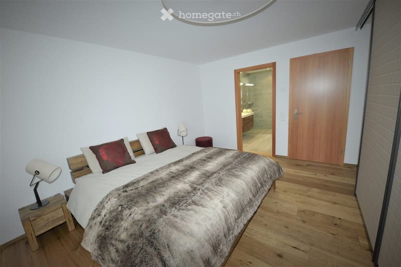 Modern bedroom with a large bed, fur throw, red pillows, wooden nightstand, and lamp. Open door leads to a bathroom with a sink and mirror.