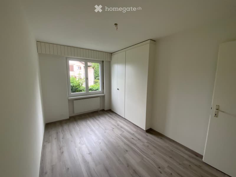 Empty room with light wooden flooring, white walls, built-in closet, window with radiator below, and a door.