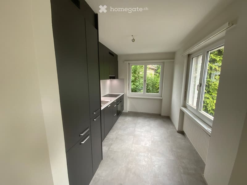 Empty kitchen with dark grey cabinets, white countertops, and large windows overlooking greenery.