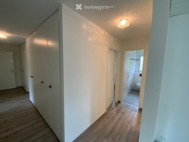 A narrow hallway with light wooden flooring, white walls, and recessed lighting. Doors lead to various rooms, including a visible bathroom with a toilet.