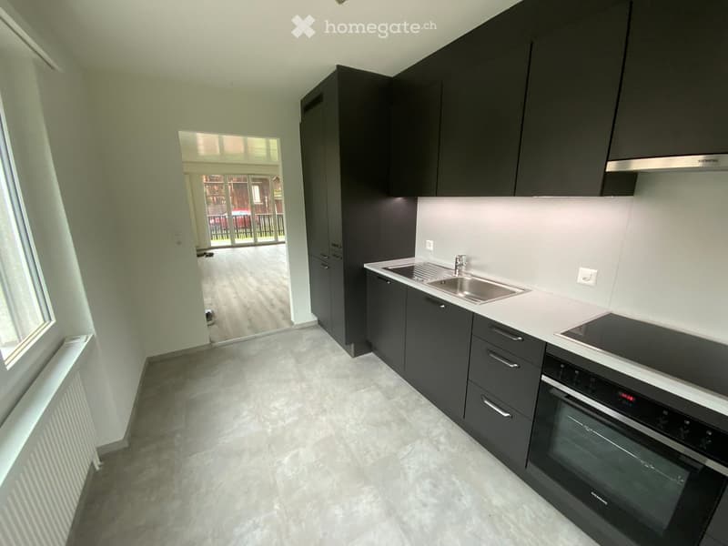 Modern kitchen with dark grey cabinets, white countertops, stainless steel sink, and built-in oven. Large window provides natural light.