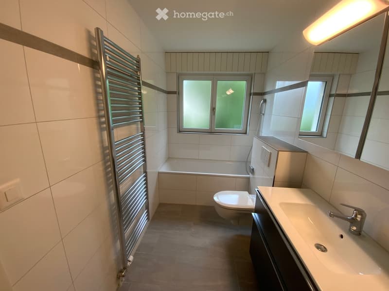 Modern bathroom with white tiles, a sink with a faucet, a toilet, a bathtub, and a radiator. The room features large windows with frosted glass and a sleek, contemporary design.
