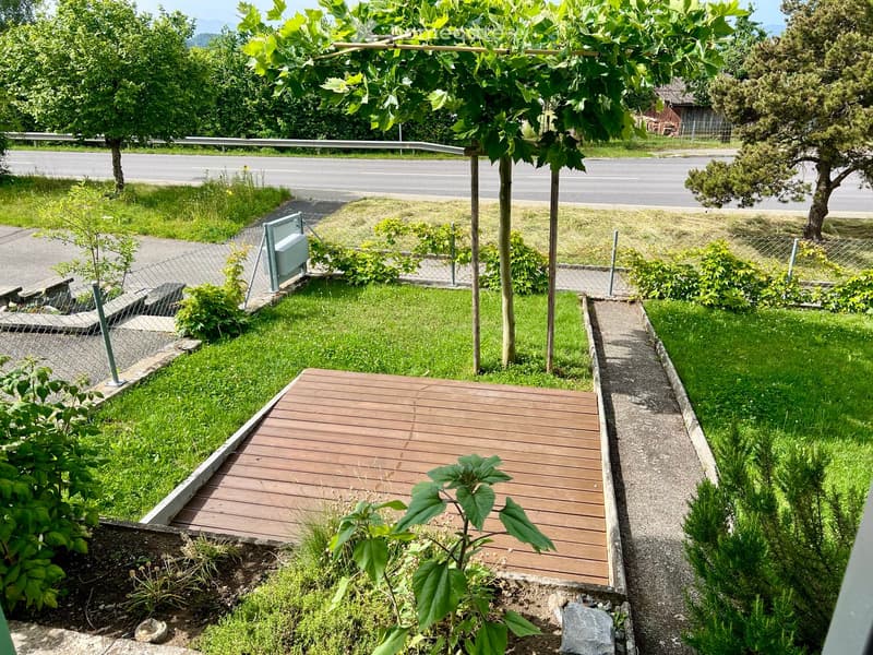 Wooden deck surrounded by green grass, small plants, and trees, with a gravel pathway and a fence along the edge.