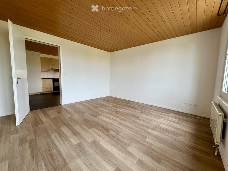 Empty room with light wooden flooring, white walls, and a wooden ceiling. An open doorway leads to a kitchen area with cabinets and appliances.