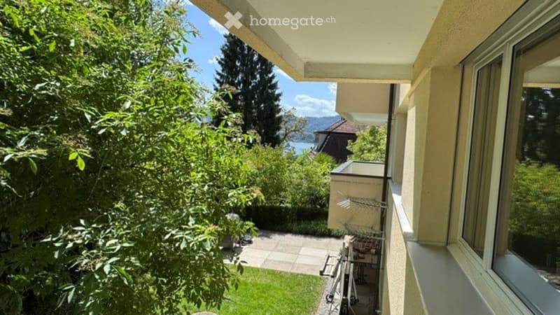 Balcony overlooking a lush garden and scenic lake view, with a paved patio and outdoor furniture visible below.