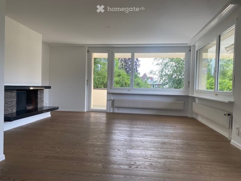 Empty room with wooden flooring, white walls, large windows, and a fireplace on one side.
