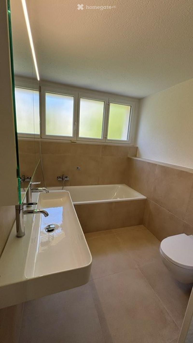 Modern bathroom with a large sink, bathtub, and toilet. The walls are tiled in beige, and there is a large mirror above the sink.