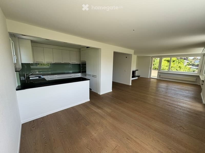 Open-plan unfurnished space with a modern kitchen area featuring white cabinets, a black countertop, and a built-in oven. The living area has wooden flooring and large windows with a view of greenery.