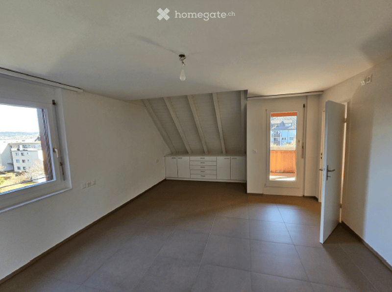 Empty attic room with exposed wooden beams, large window, built-in cabinets, and a door leading to a balcony.