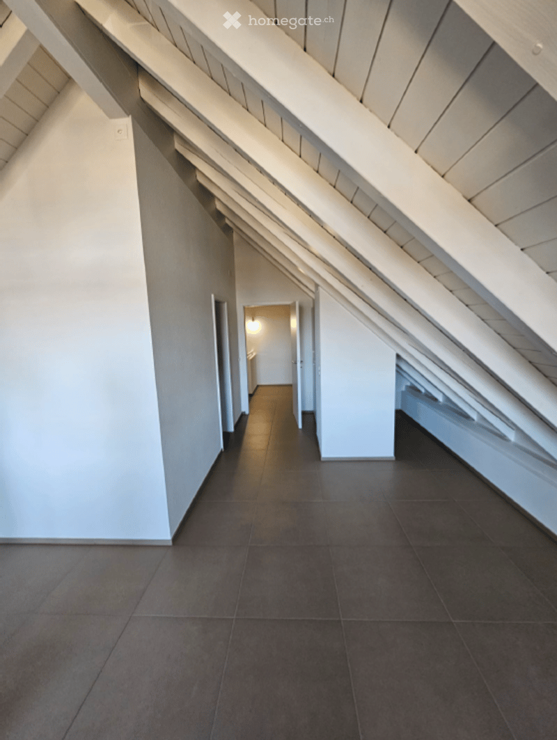 A narrow, modern hallway with white walls, dark tiled flooring, and exposed wooden beams on the ceiling. The hallway leads to a lit room at the end.