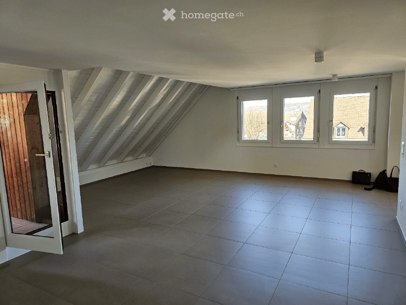 Empty attic room with slanted ceilings, large windows, tiled floor, and a wooden door leading outside.