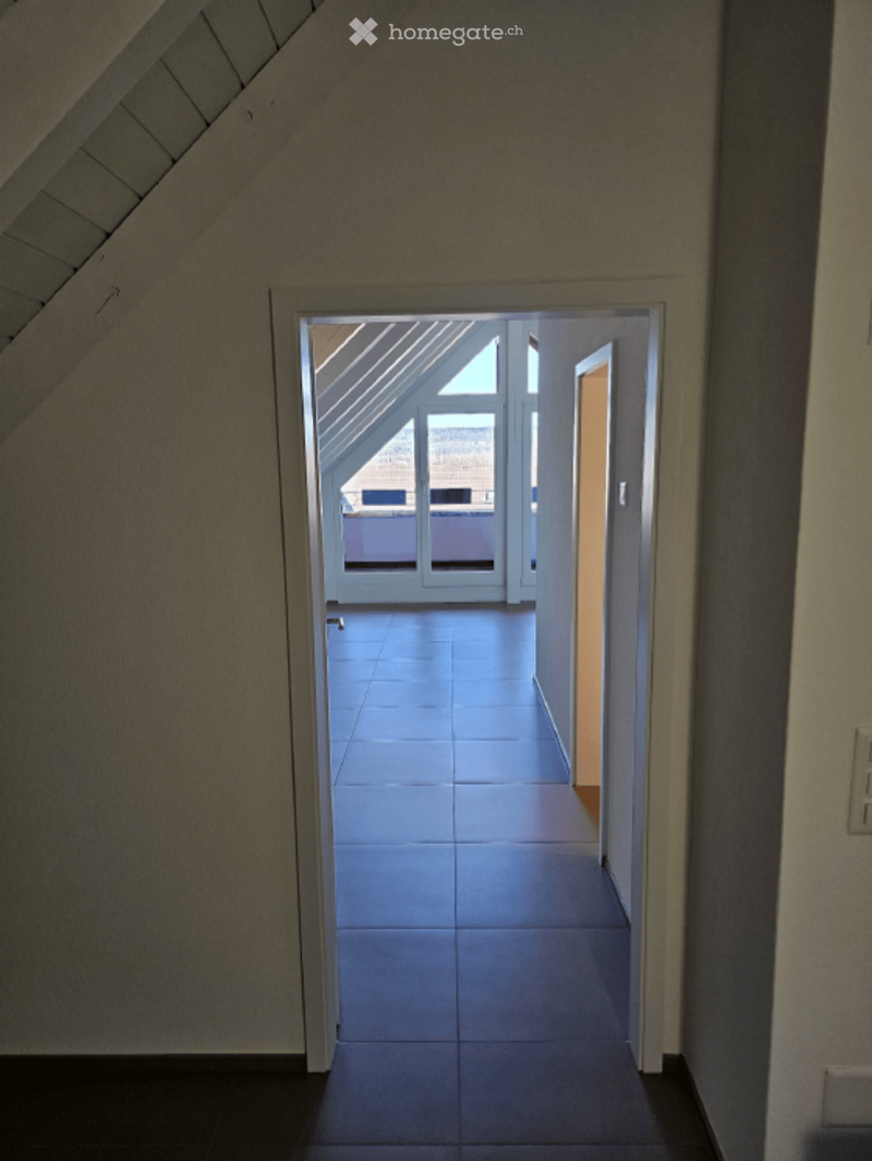A narrow hallway with a slanted ceiling leads to a bright, open space with large windows and a tiled floor.