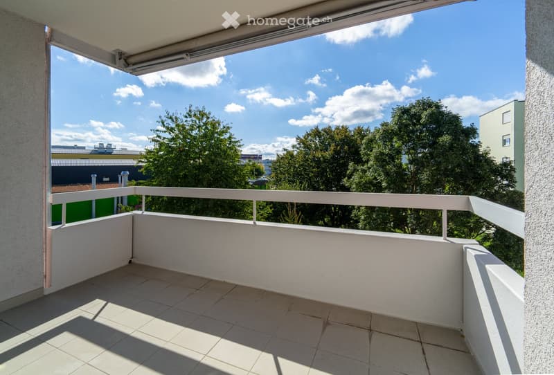 Balcony with white tiled floor, white railing, and retractable awning, overlooking a green area with trees and buildings.