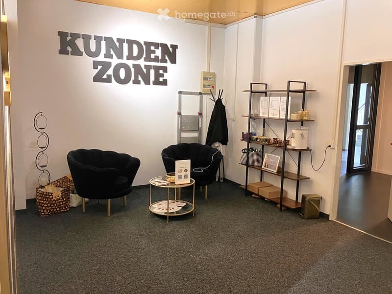 Customer zone with two black chairs, a small round table, shelves with cups and framed picture, ladder, coat, and handbag.