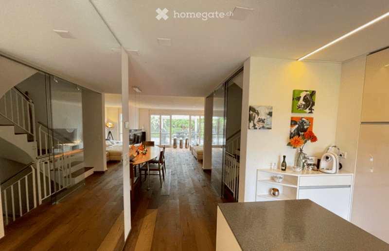 Open-plan living and dining area with wooden flooring, modern furniture including a dining table with chairs, a sofa, and a kitchen counter with a vase of flowers and framed artwork on the wall.
