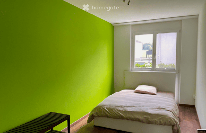 Bright bedroom with green accent wall, single bed with white bedding, small nightstand, and wooden stool near the bed.