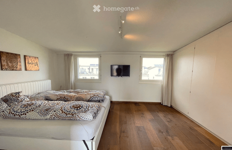 Bright bedroom with wooden flooring, large windows, white bed with patterned bedding, mounted TV, and two wall art pieces.
