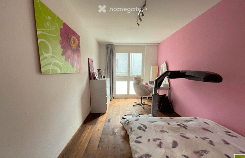 Cozy bedroom with wooden flooring, pink accent wall, large window with balcony access, bed with floral bedding, desk with chair, and wall-mounted lamp.
