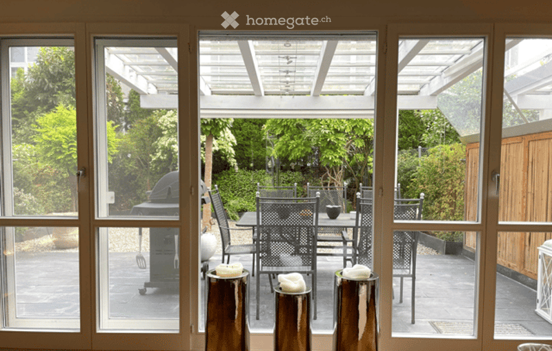 Large open patio with a dining table and chairs, surrounded by lush greenery and a pergola roof.